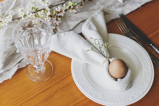 Easter egg in bunny napkin on plate with cutlery, bunny, spring flowers and rustic cloth on wooden table. Stylish Easter brunch table setting. Holiday arrangement and eco friendly decorations - Foto, imagen