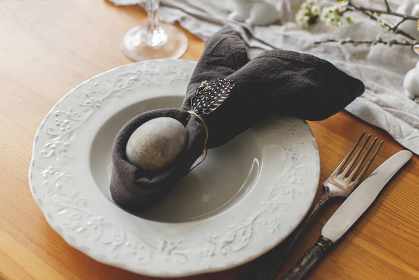 Κομψό πασχαλινό τραπέζι. Πασχαλινό αυγό σε πετσέτα λαγουδάκι στο πιάτο με μαχαιροπήρουνα, λαγουδάκι, φτερό και ρουστίκ ύφασμα σε ξύλινο τραπέζι. Πασχαλινή διάταξη τραπεζιού και οικολογική διακόσμηση - Φωτογραφία, εικόνα