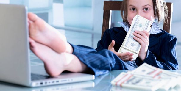 Gelukkig glimlachend jong mooi zakenmeisje zet haar voeten op een tafel en ruikt stapels US Dollar biljetten. - Foto, afbeelding