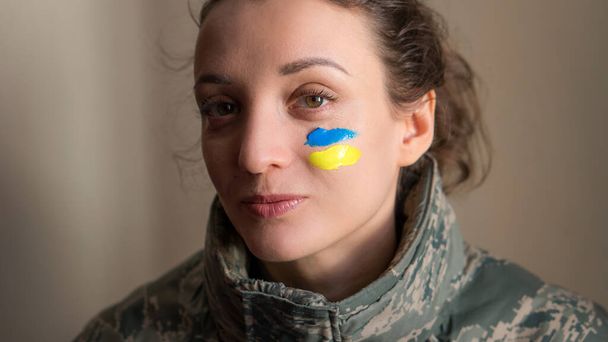 Indoor portrait of young girl with blue and yellow ukrainian flag on her cheek wearing military uniform, mandatory conscription in Ukraine, equality concepts - Foto, Imagen