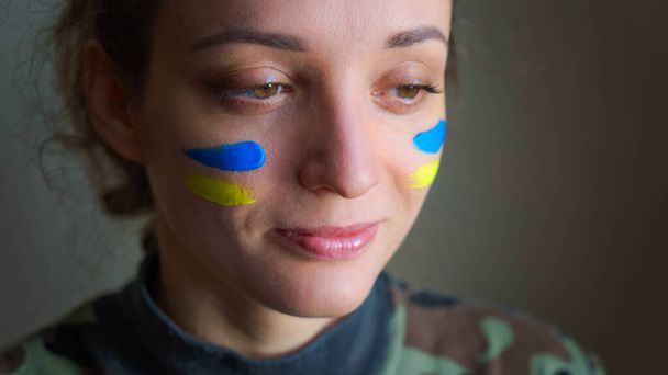 Retrato interior de niña con bandera ucraniana azul y amarilla en la mejilla con uniforme militar, reclutamiento obligatorio en Ucrania, conceptos de igualdad - Foto, imagen
