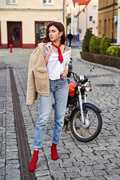 Young beautiful woman in red knitted fabric block heel sock shoes, blue denim jeans pants and teddy jacket coat posing near a motorcycle on a street - Fotoğraf, Görsel