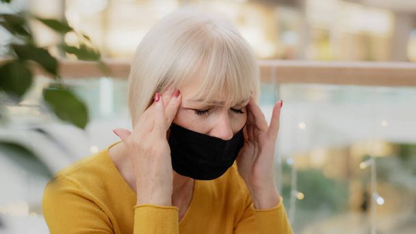 Close-up unhappy caucasian middle-aged woman in medical mask sitting indoors holding hands behind head suffering headache closing eyes feeling tired painful sensations chronic migraine health problems - Fotó, kép