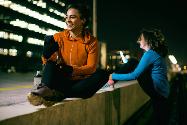 Uma esportista sorrindo se encaixa sentada lá fora à noite, descansando e usando telefone. - Foto, Imagem