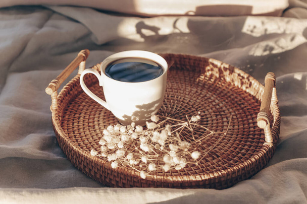Cup of americano on wicker tray in bed with grey linen bed sheet. Morning coffee concept. Sunday loneliness relaxing, hygge mood. Minimalism composition with pastel neutral colors. Light and shadow. - Photo, Image