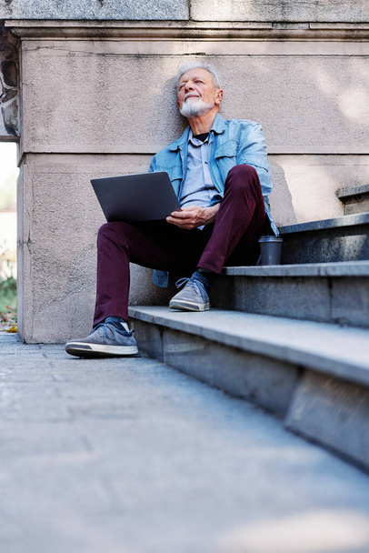 Ένας ανώτερος μαθητής κάθεται στις σκάλες του κτιρίου του πανεπιστημίου και χρησιμοποιεί το laptop του για να εξετάσει τις σημειώσεις πριν από μια εξέταση. - Φωτογραφία, εικόνα