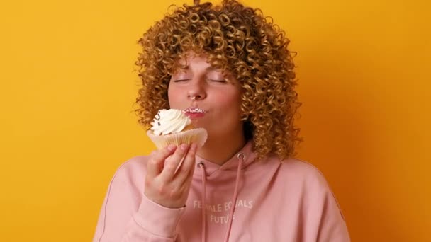 Atractiva mujer joven de 30 años con peinado afro rizos tiene expresión alegre comer pastel aislado sobre fondo amarillo de la pared - Imágenes, Vídeo