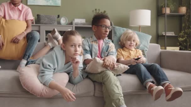Lento de cuatro niños de edad elemental diversos sentados en el sofá frente a la televisión juntos en el tiempo libre - Metraje, vídeo