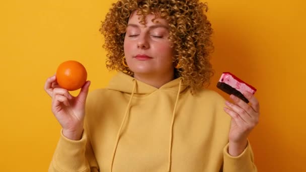 Attractive 30s woman made wrong choice between cake and fresh orange isolated over yellow. Dieting and calories concept - Footage, Video