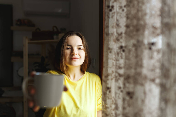 Young woman standing next to window at home and drinking coffee. Casual style indoor shoot. Morning coffee woman daily routine - Photo, Image