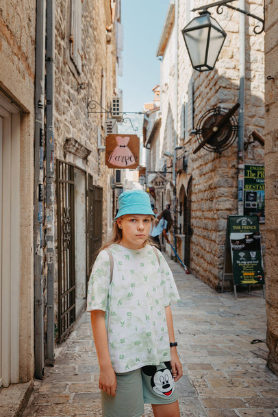 Girl Tourist Walking Through Ancient Narrow Street On A Beautiful Summer Day In Mediterranean Medieval City, Old Town Budva, Montenegro. Young Beautiful Cheerful Woman Walking On Old Street At - Zdjęcie, obraz