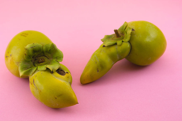 Two green persimmons with deformations that disfigure their appearance and turn them into waste for the industry on a pink background - Photo, Image