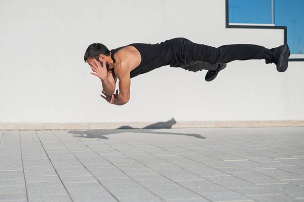 Un hombre en ropa deportiva negra salta mientras hace flexiones al aire libre. - Foto, imagen
