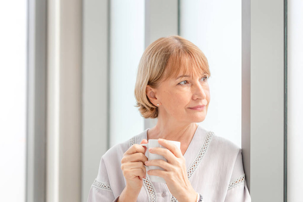 Kahve molası sırasında yeni evde rahatlatıcı kadın portresi, elinde kahve bardaklarıyla pencerenin yanında duran kadın. - Fotoğraf, Görsel