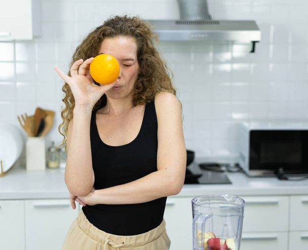smart vegetarian woman happy funny moment by hiding and closing eye behind orange while cooking fruit juice in kitchen. beautiful caucasian woman cooking healthy diet fruit or vegetable in kitchen - Foto, afbeelding