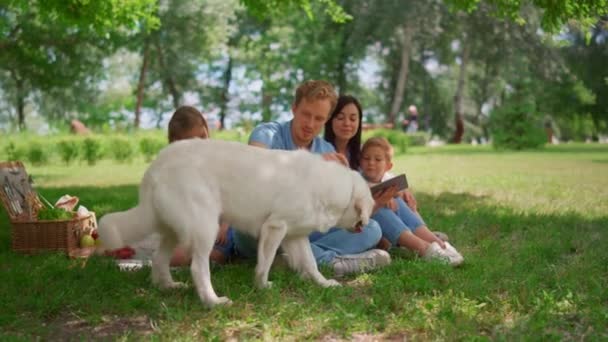 Witte hond liggen in de buurt van familie holding tablet op picknick. Vader strelen labrador. - Video