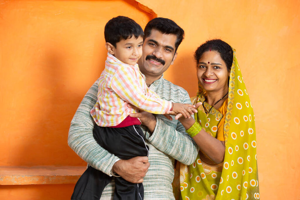 Χαρούμενη νεαρή αγροτική οικογένεια Ινδιάνων να γελάει. πατέρας κρατώντας μικρό παιδί αγόρι και μητέρα φορώντας σάρι χαμογελώντας, ζευγάρι φορώντας παραδοσιακά ρούχα με παιδί. - Φωτογραφία, εικόνα