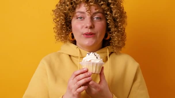 Atractiva mujer joven de 30 años con peinado afro rizos tiene expresión alegre celebración de pastel aislado sobre fondo amarillo de la pared - Imágenes, Vídeo