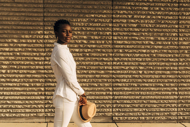 Elegante mujer afroamericana sosteniendo un sombrero mientras posa mirando a la cámara. - Foto, imagen