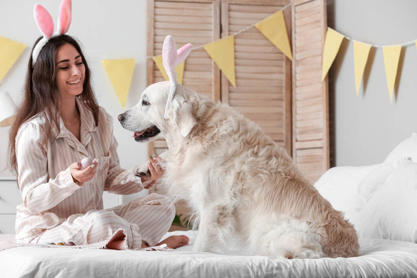 Mujer joven y adorable perro con orejas de conejo y huevos de Pascua en la cama en casa - Foto, imagen