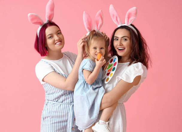 Young lesbian couple with little daughter, Easter egg and paints on color background - Photo, Image