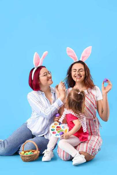 Junges lesbisches Paar mit kleiner Tochter, Ostereiern, Pinseln und Farben auf farbigem Hintergrund - Foto, Bild