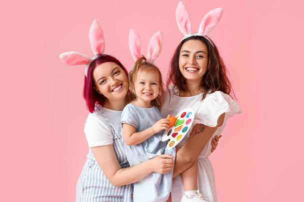 Pareja joven lesbiana con hija pequeña, huevo de Pascua y pinturas sobre fondo de color - Foto, Imagen