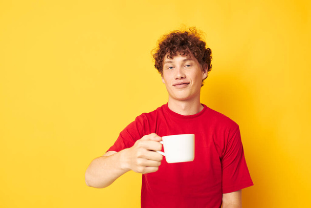 portrait of a young curly man posing with a white mug and in the hands of a drink yellow background unaltered - Fotó, kép