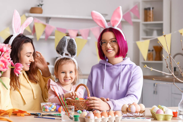 Jeune couple lesbienne avec petite fille pendant la peinture des œufs de Pâques dans la cuisine - Photo, image
