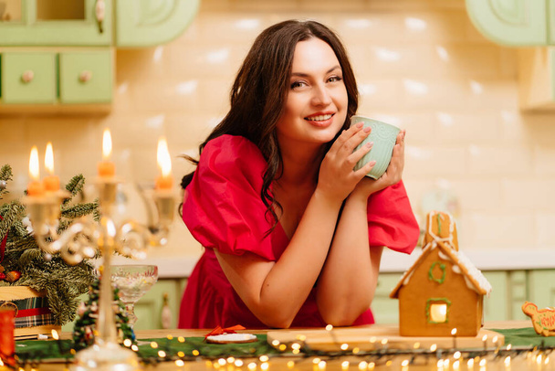 a woman in a red dress drinks from a cup in the kitchen with a gingerbread house - Фото, зображення
