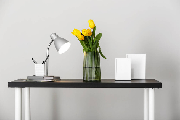 Jarrón con tulipanes, lámpara brillante y artículos de papelería en la mesa cerca de la pared de luz - Foto, Imagen