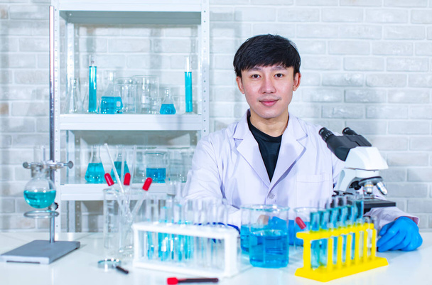 Muotokuva studio laukaus Aasian nuori ammatillinen mies tiedemies valkoinen lab takki istuu hymyillen katso kameraa takana työpöydän täynnä koeputkia dekantterilasipulloja näyte ja mikroskooppi. - Valokuva, kuva
