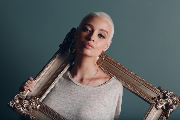 Millenial young woman with short blonde hair holds gilded picture frame in hands behind her face portrait. - Фото, зображення