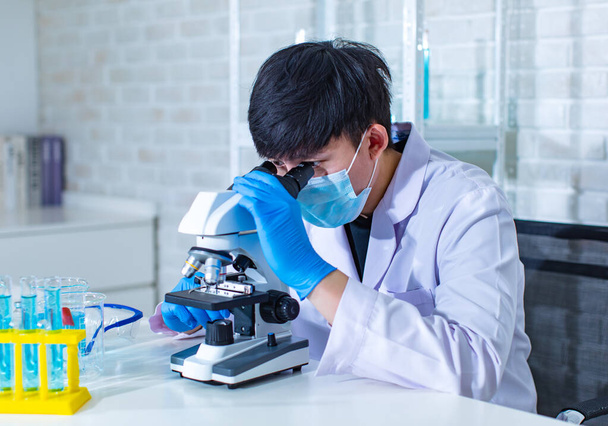 Lähikuva kasvojen kuva Aasian nuori ammatillinen mies tiedemies valkoinen lab takki yllään turvallisuus suojalasit lasit käyttäen mikroskooppi linssi etsii zoomaus mikrobiologia näyte lasilevyllä. - Valokuva, kuva