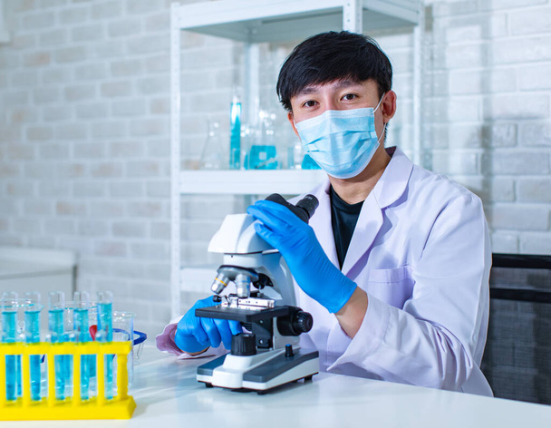 Gros plan du visage du jeune scientifique professionnel asiatique en blouse de laboratoire blanche portant des lunettes de protection de sécurité à l'aide d'une lentille de microscope regardant un échantillon de microbiologie sur une plaque de verre. - Photo, image