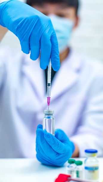 Close-up shot injectienaald zuigt coronavirus covid19 vaccin uit glazen flacon voor injectie door Aziatische professionele mannelijke wetenschapper in witte labjas met gezichtsmasker op een wazige achtergrond. - Foto, afbeelding