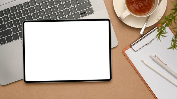 Вид зверху, Сучасний робочий простір з цифровим планшетом білий екран макет над ноутбуком, кишеньковий папір і чашка чаю на коричневому фоні
. - Фото, зображення