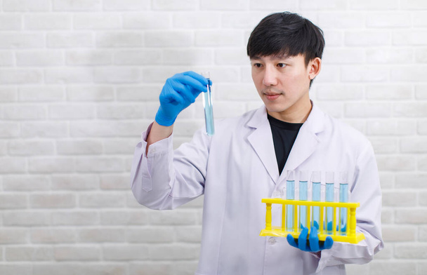 Portrait shot Asian professional male scientist in white lab coat rubber gloves and safety goggles stand smiling look at camera holding showing sample test tube rack in hands on brick wall background. - Photo, Image
