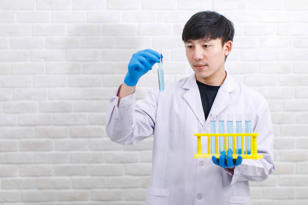 Portraitaufnahme asiatischer männlicher Wissenschaftler in weißem Labormantel, Gummihandschuhen und Schutzbrille stehen lächelnd vor der Kamera und zeigen das Probenröhrchen in der Hand auf Backsteinwand-Hintergrund. - Foto, Bild