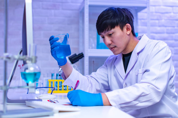 Portret studio strzał Azji profesjonalny męski naukowiec w białym fartuchu laboratorium gumowe rękawiczki siedzi uśmiechnięty patrzeć na aparat trzymając próbkę probówki w rękach w szpitalnym laboratorium pracowni. - Zdjęcie, obraz
