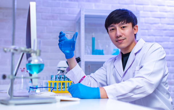 Portrait-Studioaufnahme eines asiatischen männlichen Wissenschaftlers in weißem Labormantel mit Gummihandschuhen, der lächelnd in die Kamera blickt, die das Probenröhrchen in der Hand im Arbeitszimmer des Krankenhauslabors zeigt. - Foto, Bild