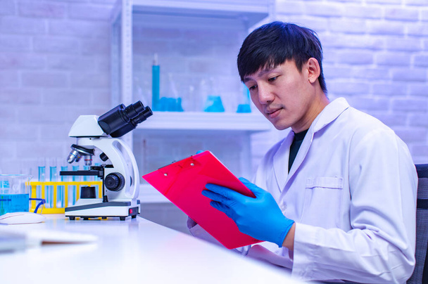 Portrait studio shot of Asian professional male scientist in white lab coat rubber gloves sitting smiling look at camera holding showing sample test tube in hands in hospital laboratory working room. - Photo, Image
