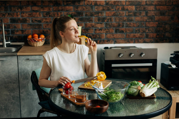 Young woman preparing vegetarian greens salad at home and tasting yellow pepper - 写真・画像