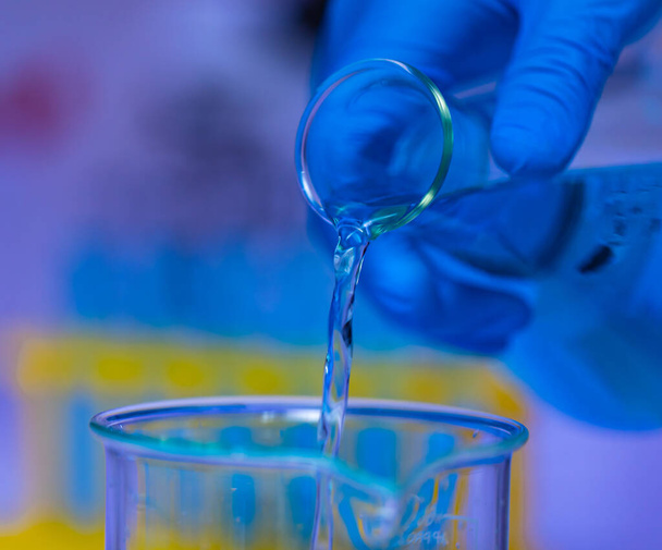 Primer plano de estudio de solución azul reactivo muestra líquida vertiendo de Erlenmeyer frasco en vaso de precipitados de vidrio por mano científica usando guantes de goma de seguridad en frente de fondo borroso tubo de ensayo rack. - Foto, imagen