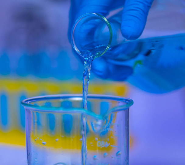 Closeup studio shot of blue solution reagent liquid sample pouring from Erlenmeyer flask into glass beaker by scientist hand wearing safety rubber gloves in front of blurred test tube rack background. - Foto, Imagem