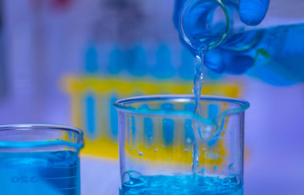 Lähikuva studio laukaus sinisen liuoksen reagenssinestenäyte kaatamalla Erlenmeyer pullosta lasi dekantterilasiin tiedemies käsin käyttäen turvallisuus kumi käsineet edessä hämärtynyt koeputki teline tausta. - Valokuva, kuva