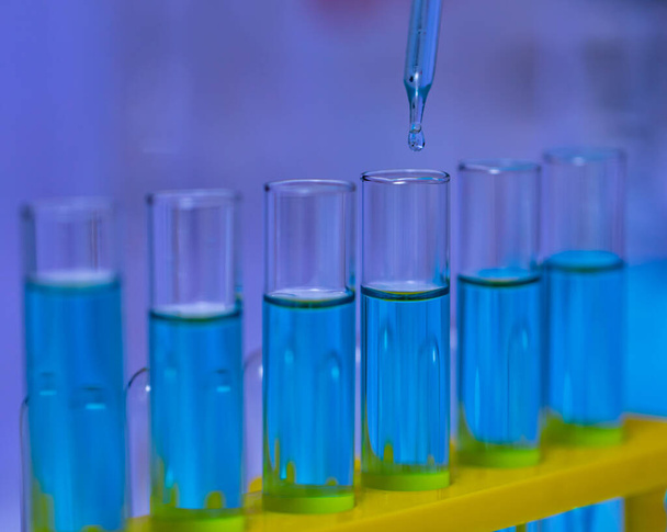 病院研究室での科学実験を分析するためにラック内のガラスウェア試験管ラインに青色の試薬溶剤溶液液サンプルをドロップ科学研究室のガラスドロッパーの閉鎖スタジオショット. - 写真・画像