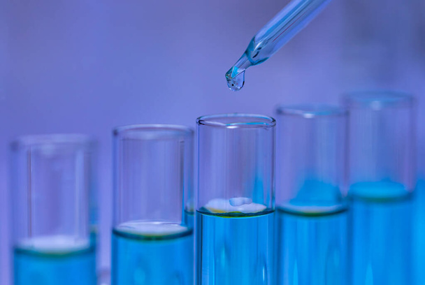 Nahaufnahme Studioaufnahme eines wissenschaftlichen Laborglastropfers, der eine Probe mit blauem Reagenzlösungsmittel in ein Reagenzglas fallen lässt, um wissenschaftliche Experimente im Krankenhauslabor zu analysieren. - Foto, Bild