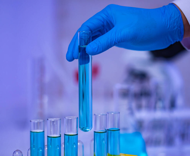Close-up studio shot van wetenschapper hand dragen rubberen handschoenen oppakken blauw reagens oplosmiddel vloeistof monster in glazen reageerbuis van gele kunststof rek in laboratorium met effect licht vlam. - Foto, afbeelding