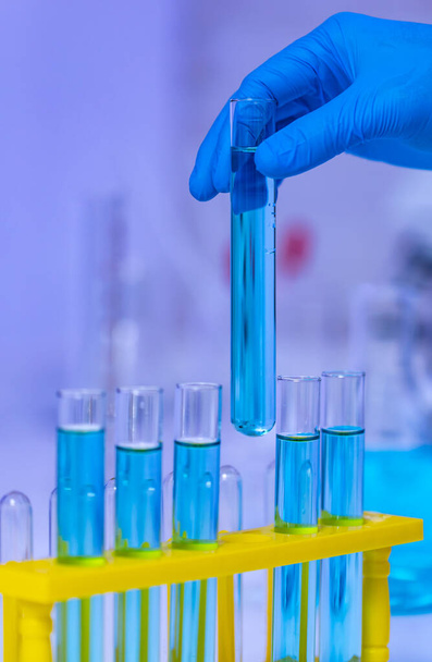Close-up studio shot van wetenschapper hand dragen rubberen handschoenen oppakken blauw reagens oplosmiddel vloeistof monster in glazen reageerbuis van gele kunststof rek in laboratorium met effect licht vlam. - Foto, afbeelding